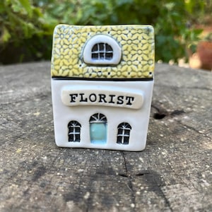 May include: A ceramic miniature house with a yellow floral patterned roof and the word "FLORIST" on the front. The house has three arched windows and a small arched window on the roof. The background is blurred green foliage.