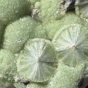 Pode incluir: Um aglomerado de cristais minerais verde-lima pálido com formações radiais distintas em forma de leque e uma textura fibrosa e cintilante. O espécime apresenta uma combinação de superfícies arredondadas e granulares com estruturas cristalinas radiantes.