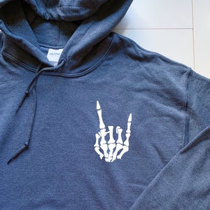 Puede incluir: Una sudadera con capucha azul oscuro con una mano de esqueleto blanca haciendo el gesto de rock and roll.