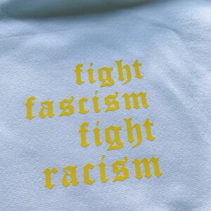 Puede incluir: Una sudadera blanca con las palabras "fight fascism fight racism" impresas en amarillo en la parte delantera.