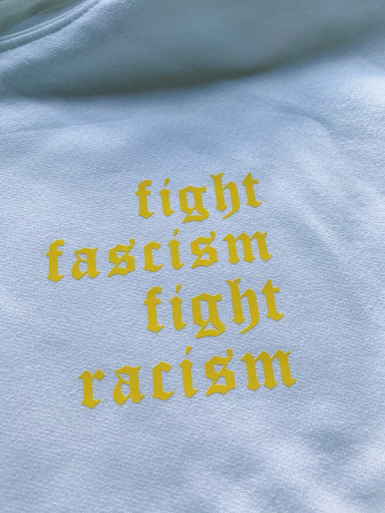 Puede incluir: Una sudadera blanca con las palabras "fight fascism fight racism" impresas en amarillo en la parte delantera.