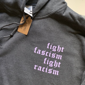 Puede incluir: Una sudadera con capucha gris oscuro con las palabras "fight fascism fight racism" impresas en morado claro en la parte delantera.