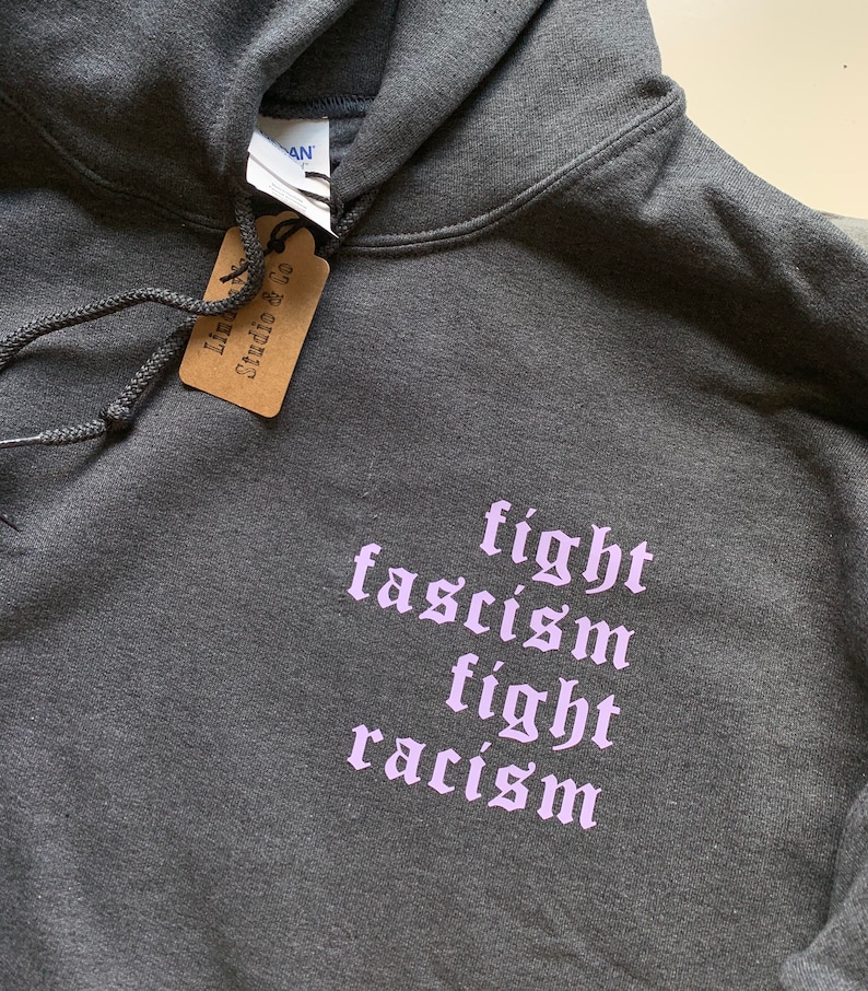 Puede incluir: Una sudadera con capucha gris oscuro con las palabras "fight fascism fight racism" impresas en morado claro en la parte delantera.