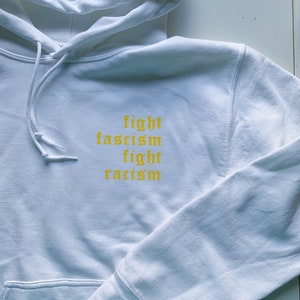 Puede incluir: Sudadera con capucha blanca con el texto "fight fascism fight racism" en amarillo.