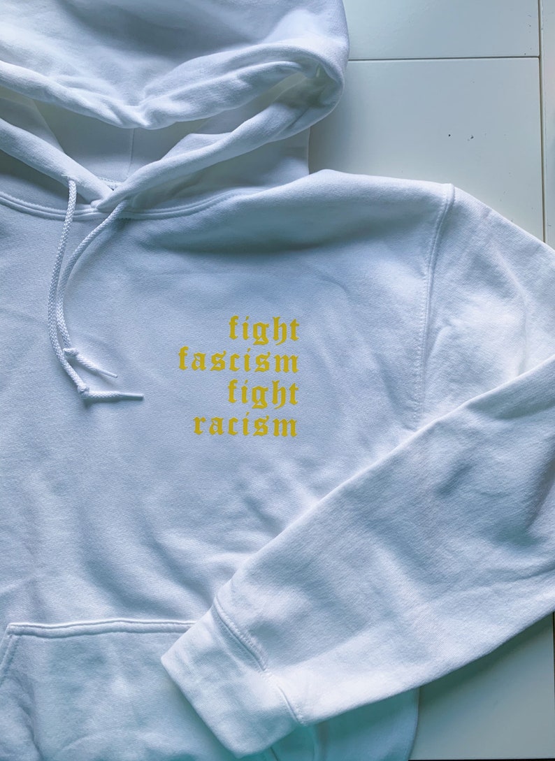 Puede incluir: Sudadera con capucha blanca con el texto "fight fascism fight racism" en amarillo.