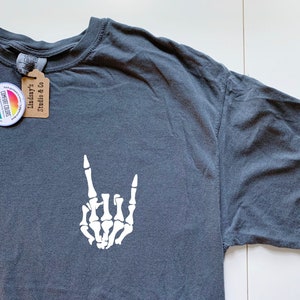Puede incluir: Una camiseta gris oscuro con una mano de esqueleto blanca haciendo el gesto de rock and roll. La camiseta tiene una etiqueta adjunta que dice "Lindsay's Studio & Co."