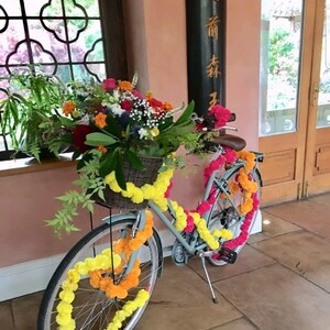 May include: A light blue bicycle decorated with yellow and orange marigold flower garlands. The bicycle has a basket filled with flowers and greenery. The bicycle is parked in front of a wooden door with a sign that reads "香  前  林".