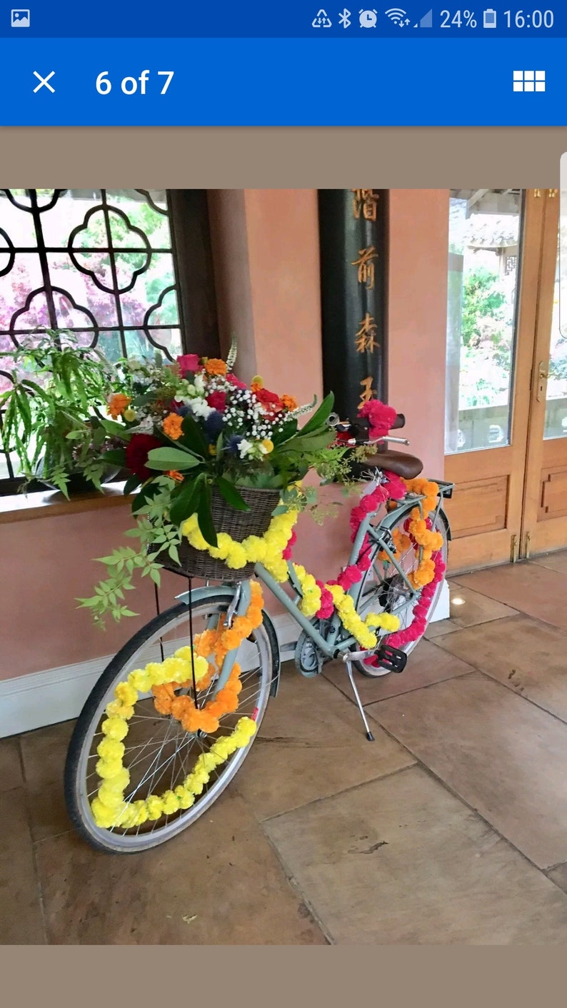May include: A light blue bicycle decorated with yellow and orange marigold flower garlands. The bicycle has a basket filled with flowers and greenery. The bicycle is parked in front of a wooden door with a sign that reads "香  前  林".