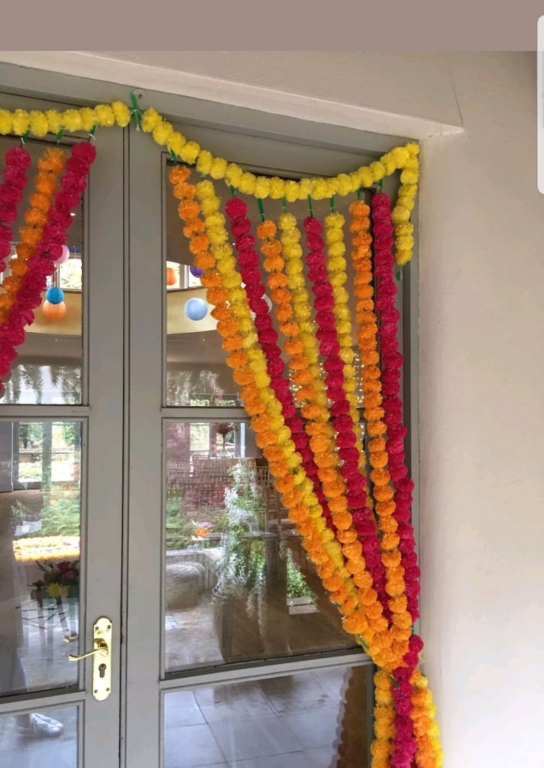 May include: A doorway decorated with a garland of yellow, orange, and pink marigold flower balls. The garland is draped over the top of the door and hangs down the sides.