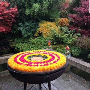May include: A circular arrangement of yellow and pink marigold flower pom-poms on a black metal stand. The stand has three legs and a ring handle. The flowers are arranged in concentric circles, with the yellow flowers on the outside and the pink flowers in the centre.