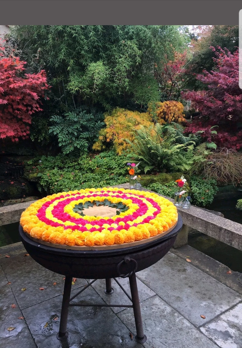 May include: A circular arrangement of yellow and pink marigold flower pom-poms on a black metal stand. The stand has three legs and a ring handle. The flowers are arranged in concentric circles, with the yellow flowers on the outside and the pink flowers in the centre.