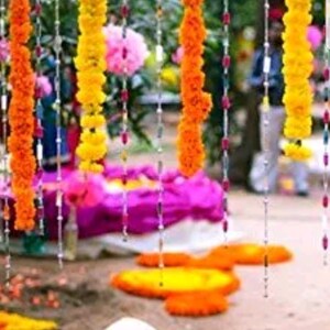 May include: A string of yellow marigold flower garlands hanging from a ceiling, with white beads strung between the flowers. The garlands are hanging in front of a purple and white fabric backdrop.