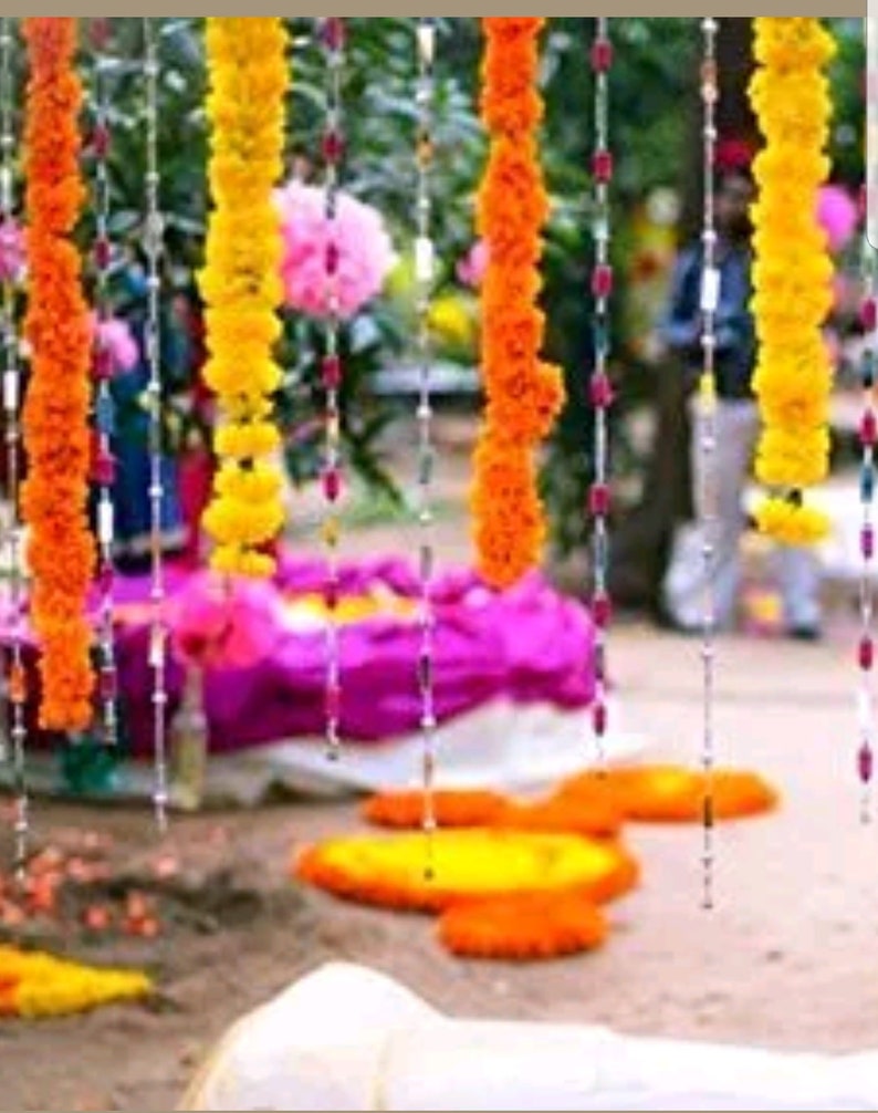May include: A string of yellow marigold flower garlands hanging from a ceiling, with white beads strung between the flowers. The garlands are hanging in front of a purple and white fabric backdrop.
