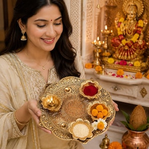 May include: A round, ornate gold-toned metal puja thali plate featuring intricate embossed patterns. The tray holds a small lit oil lamp, a bowl of bright red powder, a bowl of yellow marigold flowers, and a bowl of white rice grains.