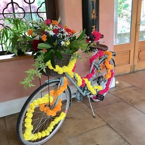 May include: A light blue bicycle decorated with yellow and pink flower garlands. The bicycle has a basket filled with flowers and is parked in front of a building with a window and a wooden door.