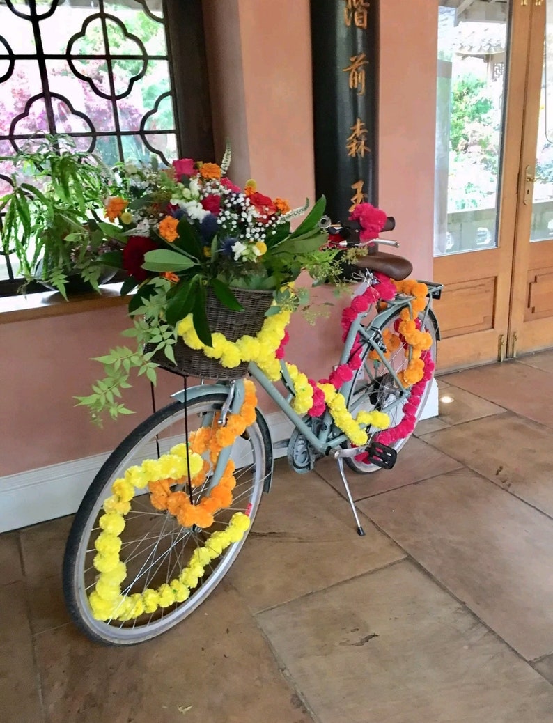 May include: A light blue bicycle decorated with yellow and pink flower garlands. The bicycle has a basket filled with flowers and is parked in front of a building with a window and a wooden door.