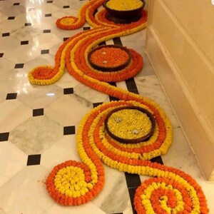 May include: A decorative floor arrangement made of yellow and orange marigold flower garlands. The garlands are arranged in a swirling pattern with small bowls filled with marigolds in the centre of each swirl.