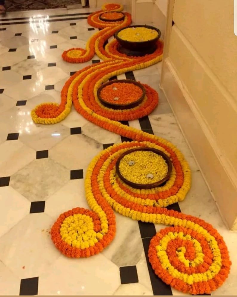 May include: A decorative floor arrangement made of yellow and orange marigold flower garlands. The garlands are arranged in a swirling pattern with small bowls filled with marigolds in the centre of each swirl.