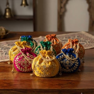 Könnte beinhalten: Eine Sammlung von Beuteltaschen in leuchtenden Farben wie Gelb, Pink, Blau, Grün und Orange. Jede Tasche ist mit aufwendigen goldenen Blumenstickereien und einem Perlenhenkel versehen. Ein kleines rechteckiges Schild trägt die Aufschrift HANDCRAFTED LUXURY.