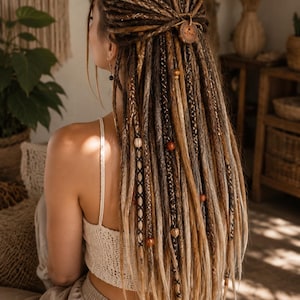 Könnte beinhalten: Ein Set aus langen, mehrfarbigen braunen und blonden synthetischen Dreadlocks, die zu einer halben Hochsteckfrisur gestylt sind. Das Haar ist mit verschiedenen dekorativen Elementen wie Holzperlen, Garnwicklungen und einem runden Holzanhänger verziert.