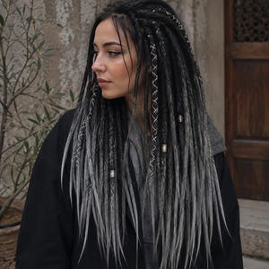 Könnte beinhalten: Lange Dreadlocks mit einem Farbverlauf von Schwarz zu Grau. Die Haarverlängerungen sind mit weißem Garn umwickelt und mit kleinen metallischen Perlen verziert, was für eine strukturierte Optik sorgt, die bis über die Schultern reicht.