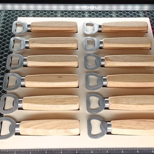 Könnte beinhalten: Zwölf Flaschenöffner aus Holz mit Metall-Flaschenöffnermechanismen. Die Flaschenöffner sind in zwei Reihen zu sechs auf einer weißen Oberfläche angeordnet.