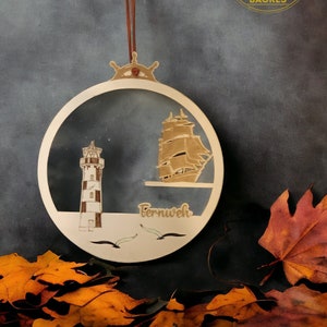 May include: A wooden round ornament with a lighthouse, a ship, and the word "Bernweh" on it. The ornament is hanging from a brown leather cord. The ornament is against a gray background.