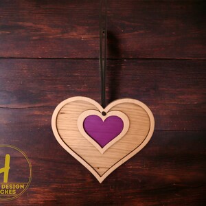 May include: Wooden heart-shaped wall decor with a layered design. The outer layer is light brown, the middle layer is a lighter shade, and the center is a vibrant purple. A black cord is attached for hanging. The logo "HOLZ DESIGN BACKES" is in the lower left.