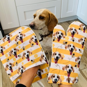 May include: A pair of yellow and white oven mitts with a pattern of a dog's face and bees. The mitts are made of fabric and have a striped design.