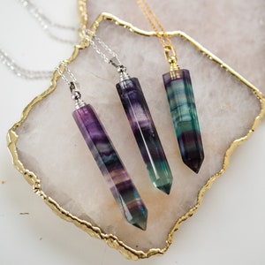 May include: Three necklaces with fluorite crystal pendants. The pendants are long and pointed, with a mix of purple, green, and blue colors. The necklaces are displayed on a white agate slice with a gold rim.