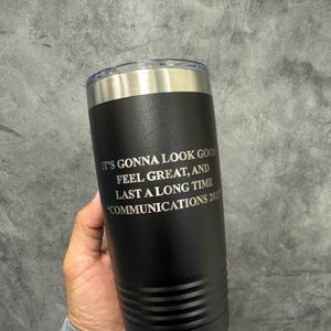 May include: Black insulated tumbler with a silver rim and lid. The tumbler has the text "IT'S GONNA LOOK GOOD FEEL GREAT, AND LAST A LONG TIME COMMUNICATIONS 2025" in white. The tumbler is cylindrical and appears to be made of stainless steel.