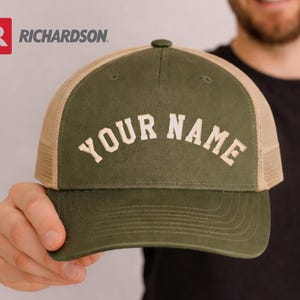 May include: A green and tan trucker hat with the text "YOUR NAME" in white block letters. The hat has a curved brim and a mesh back. The Richardson logo is visible in the upper left corner.