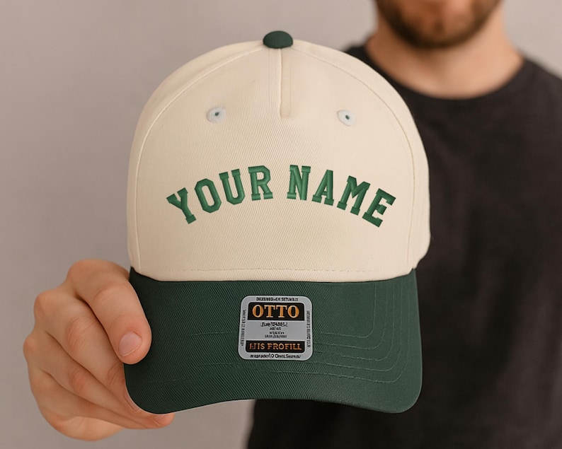 May include: Cream-colored baseball cap with a dark green brim and a green button on top. The text "YOUR NAME" is embroidered in green on the front. A small tag with text is visible on the brim.
