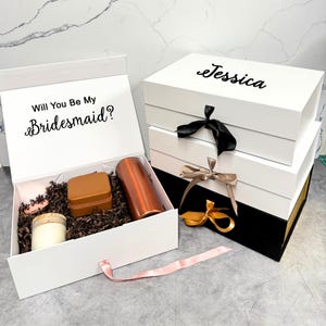 May include: White gift boxes with black and gold accents, tied with ribbons. One box is open, revealing a candle, a small box, and a copper-colored tumbler. The open box has the text "Will You Be My Bridesmaid?"