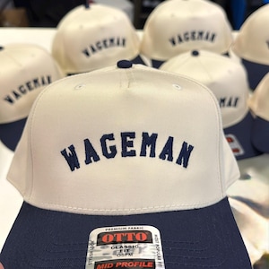 May include: A collection of baseball caps with a cream-colored crown and a navy blue brim. The word "WAGEMAN" is embroidered in navy blue on the front. The caps are made by OTTO and have a mid-profile design.