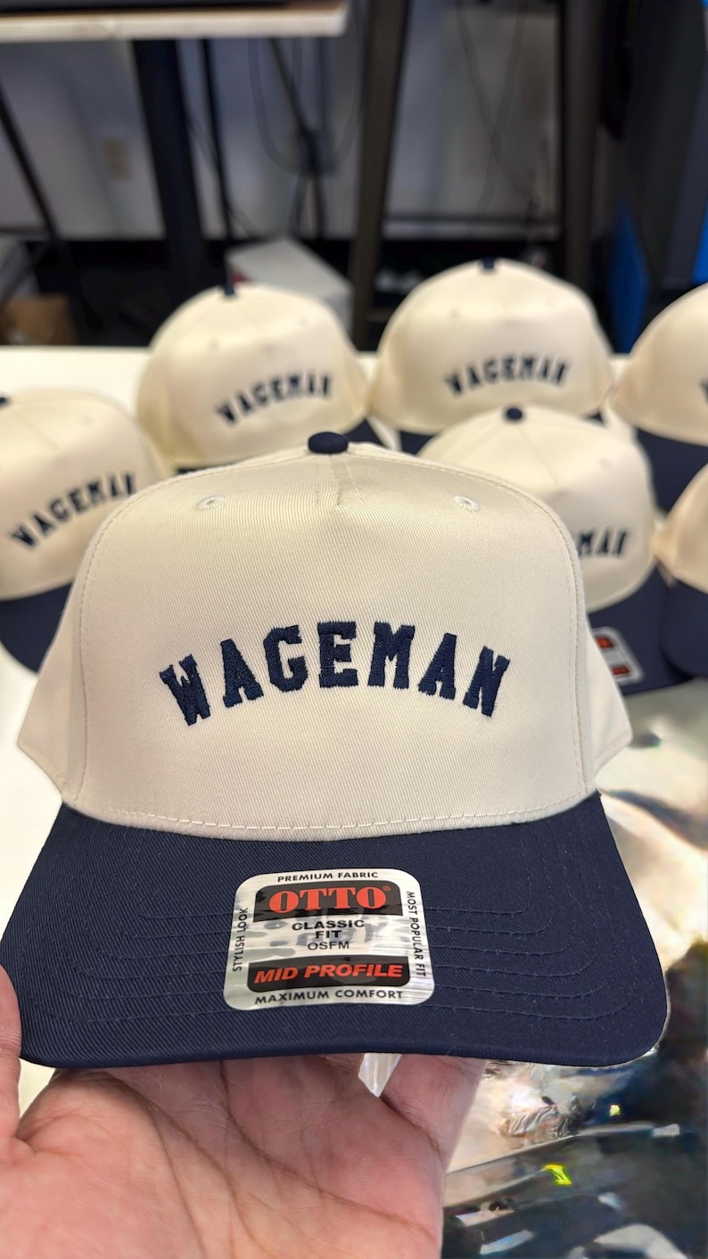 May include: A collection of baseball caps with a cream-colored crown and a navy blue brim. The word "WAGEMAN" is embroidered in navy blue on the front. The caps are made by OTTO and have a mid-profile design.