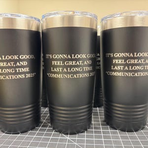 May include: Black insulated tumblers with silver rims and clear lids. Each tumbler has the text "IT'S GONNA LOOK GOOD, FEEL GREAT, AND LAST A LONG TIME COMMUNICATIONS 2025" etched in white. The tumblers are cylindrical.