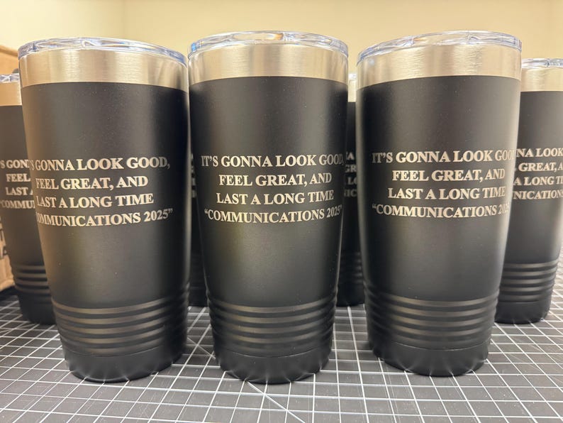 May include: Black insulated tumblers with silver rims and clear lids. Each tumbler has the text "IT'S GONNA LOOK GOOD, FEEL GREAT, AND LAST A LONG TIME COMMUNICATIONS 2025" etched in white. The tumblers are cylindrical.