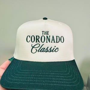 May include: A white and green baseball cap with the text "THE CORONADO Classic" embroidered in green. The cap has a dark green brim and a green button on top. The hat is made of a textured fabric.