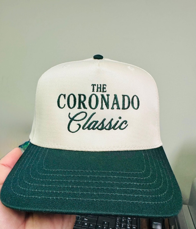 May include: A white and green baseball cap with the text "THE CORONADO Classic" embroidered in green. The cap has a dark green brim and a green button on top. The hat is made of a textured fabric.