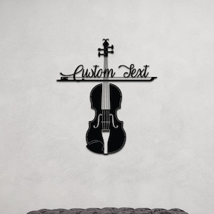 Pode incluir: Arte de parede de violino de metal preto com o texto "Custom Text" acima. O violino está pendurado em uma parede branca acima de um banco estofado cinza.