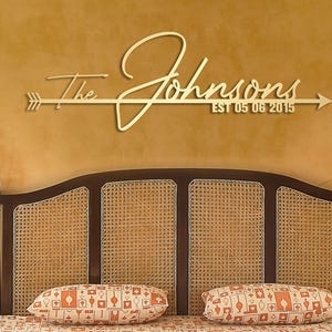 May include: A gold-coloured metal wall sign featuring an arrow design with the text The Johnsons and EST 05 06 2015. The sign is mounted horizontally on a wall above a bed frame with a woven cane headboard and two patterned pillows.