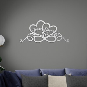 May include: A silver metal wall decoration showing two hearts intertwined, with the names "Jessica" and "David" in cursive. The design incorporates an infinity symbol and decorative swirls, ideal for interior decor.