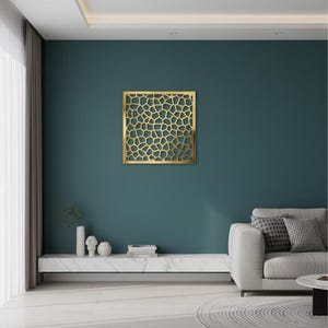 May include: A square gold-coloured metal wall panel featuring an intricate, open-work geometric pattern of irregular, interconnected polygons. The panel is mounted on a solid, dark teal wall, creating a high-contrast visual display.