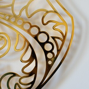 May include: A gold-colored metal cutout with a decorative design featuring swirls and circles. The cutout is in the shape of a rounded rectangle.