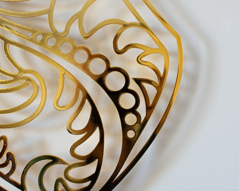 May include: A gold-colored metal cutout with a decorative design featuring swirls and circles. The cutout is in the shape of a rounded rectangle.