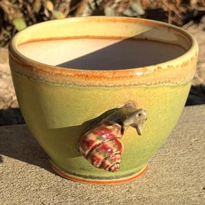 May include: A handmade ceramic bowl with a green exterior and a light brown interior. A snail sculpture with a red and brown shell is attached to the side of the bowl. The bowl has a rustic, earthy aesthetic.