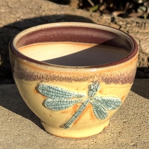 May include: A handmade ceramic bowl with a dragonfly design. The bowl features a light yellow exterior with a brown rim and a dragonfly in shades of blue and green. The bowl is likely used for serving or display.