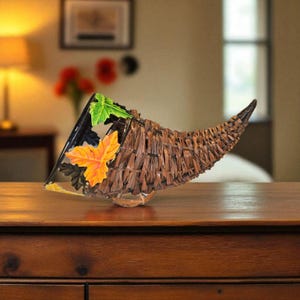 May include: A decorative cornucopia made of woven brown material, with a black base and colorful leaf accents. The horn-shaped basket is displayed on a wooden surface, suggesting a fall or harvest theme.