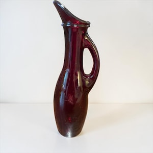 May include: A tall, ruby-red glass pitcher with a curved spout and a handle. The pitcher has a slender, elegant shape and a smooth, glossy finish. The handle is a loop, and the spout is angled upwards. The pitcher is likely for serving beverages.